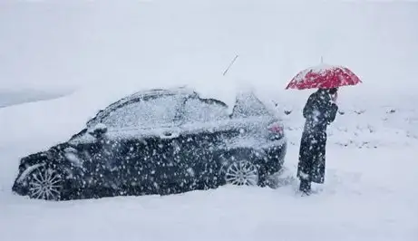  A woman stands beside her parked red car in a snowy landscape, holding an open umbrella for shelter. Snowflakes fall gently around her, and the car is lightly dusted with snow, suggesting cold winter conditions.