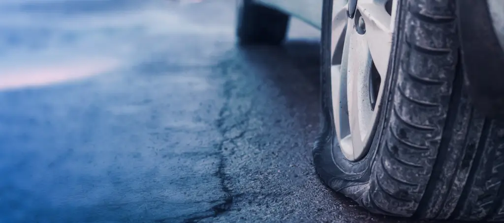 Close-up of a car’s rear tire with a visible puncture, showing a nail embedded in the tread and slight deflation. The vehicle is parked on the roadside with hazard lights on.